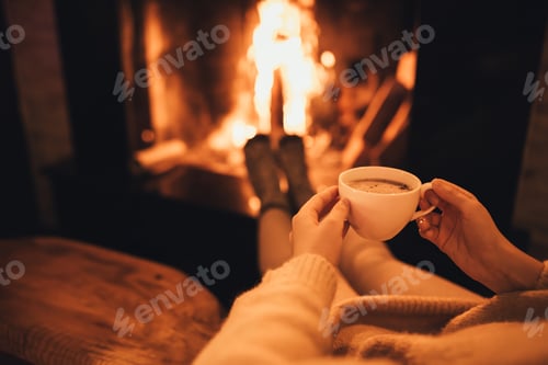 Preview: Woman drinkink hot cocoa sitting by the fire in front of cozy fireplace.