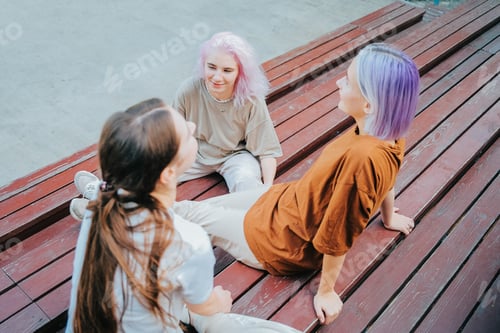 Preview: Modern teenage girls with colorful dyed hair sitting on bench in park. Women chatting, gossiping and