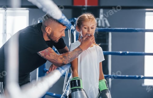 Preview: Standing on the boxing ring. Young tattooed boxing coach teaches cute little girl in the gym