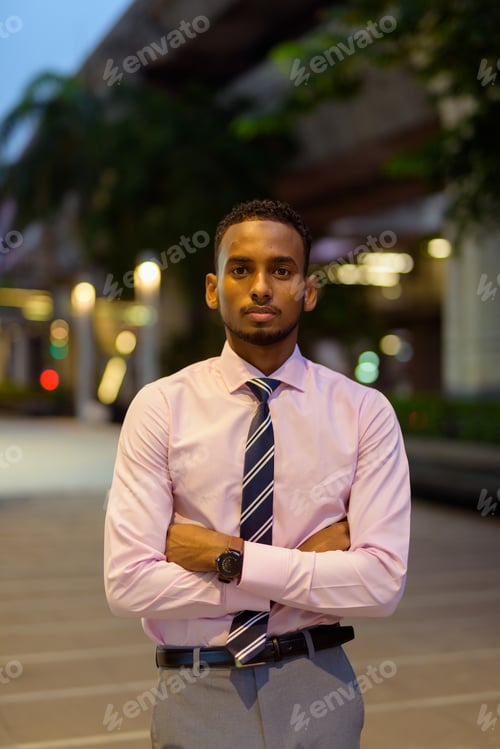 Preview: Young African businessman outdoors in city at night with arms crossed