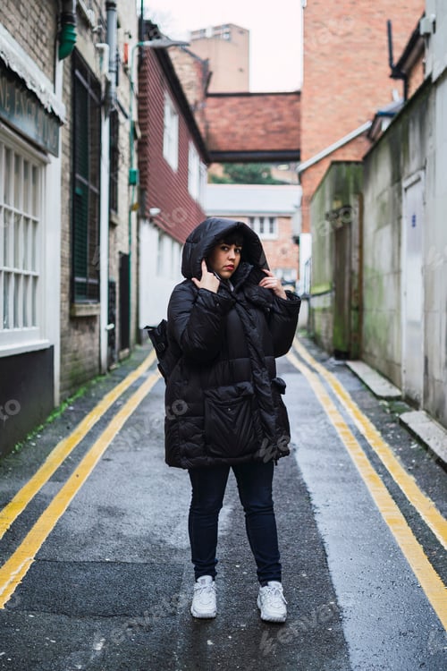 Preview: Vertical shot of a young Caucasian female with a black coat posing in an old neighborhood