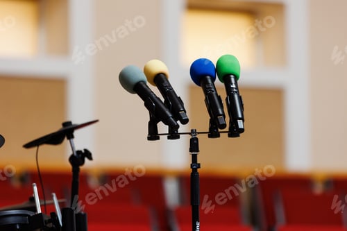 Preview: Four coloured microphones on stand on podium and a lot of red chairs blurred in the background. C