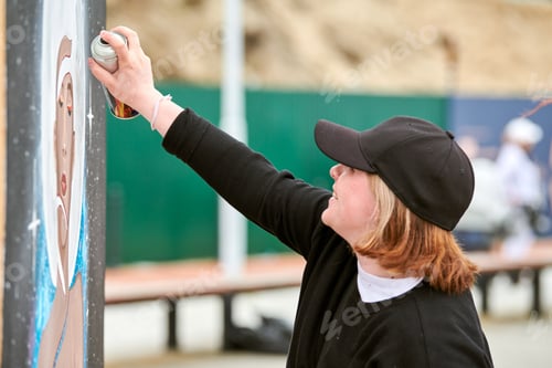 Preview: Female artist in black cap is painting picture with paint spray can spraying it onto canvas