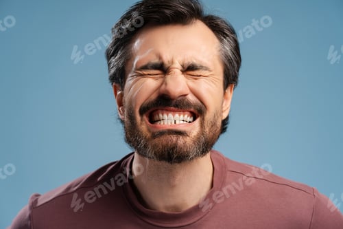Preview: Young man screw up his eyes while posing in studio and showing his tooth, isolated on blue