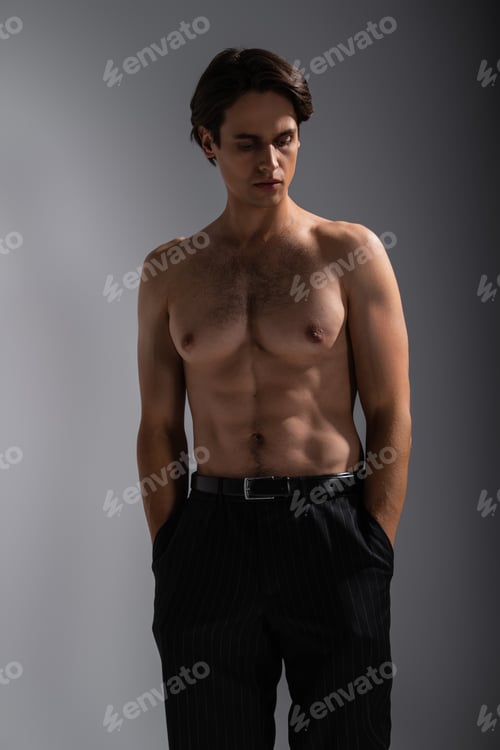 Preview: good looking man with muscular body posing with hands in pockets of black trousers on grey