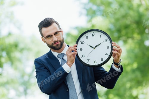 Preview: young businessman holding clock and looking at camera while standing in park