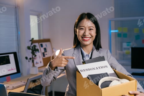 Preview: Young office worker carrying personal belongings in a box and pointing at her resignation letter whi