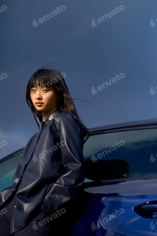 Preview: Woman in leather jacket leans against a blue car under a cloudy sky. Brandenburg, Germany