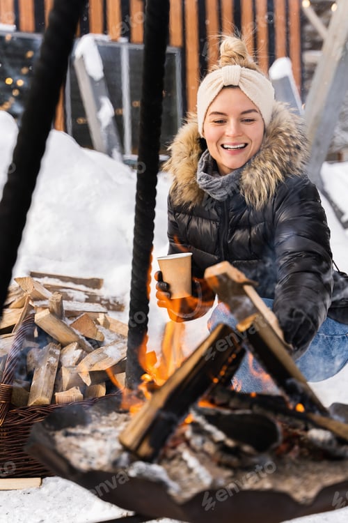 Preview: Beautiful woman warming up by the fire pit during cold winter day