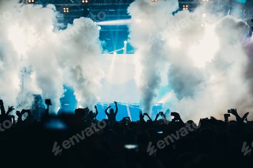 Preview: Stage lights and crowd of audience with hands raised at a music festival.
