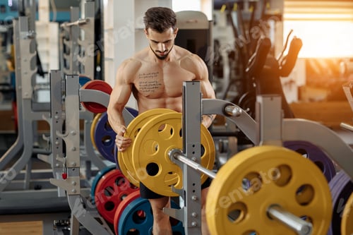 Preview: Strong athletic young man bodybuilder fitness model put weights on barbell on the bench in gym.