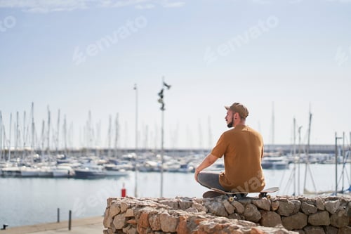 Preview: Man in cap relaxed sitting on a skateboard looking out over the harbor