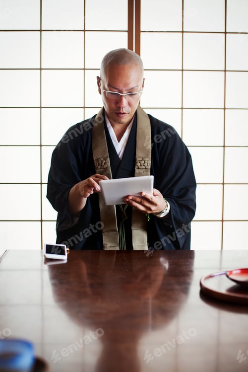 Preview: Buddhist monk with shaved head wearing black robe sitting indoors at a table, using digital tablet.