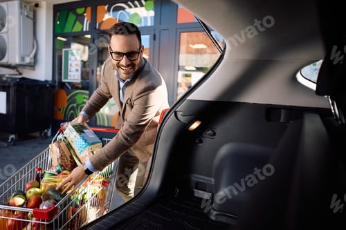 Preview: Happy man with shopping cart packing groceries into car trunk in front of the store.