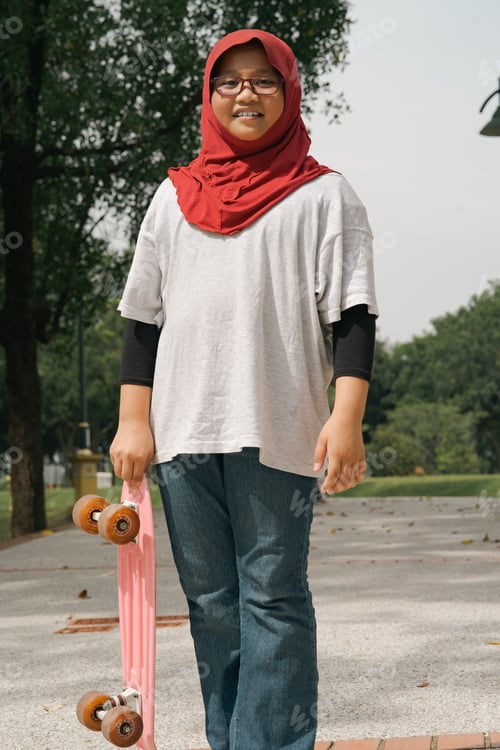 Preview: Young girl wearing hijab is standing while holding pennyboard in the park.