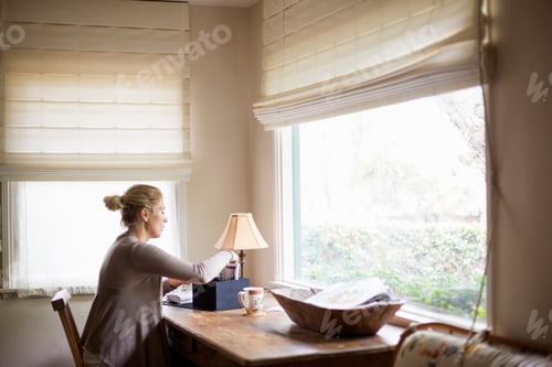 Preview: Blond woman sitting at a desk by a window, sorting through a box with photographs.