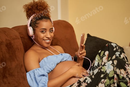 Preview: pregnant African woman on couch while listening to music with headphones looking at the camera