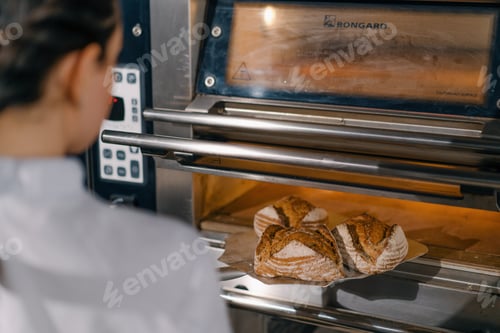 Preview: female baker takes out freshly baked fresh bread from the oven and puts it on shelf in kitchen