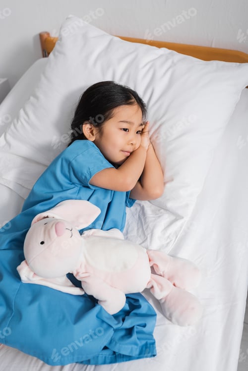 Preview: high angle view of dreamy asian child lying on hospital bed near toy bunny and looking away