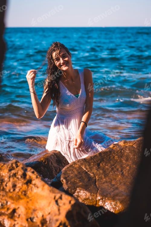 Preview: A young Caucasian woman in a wet white dress sitting on a rock