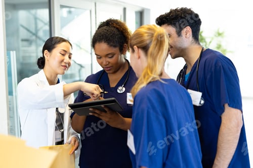 Preview: Four happy diverse healthcare colleagues looking at tablet together in hospital corridor