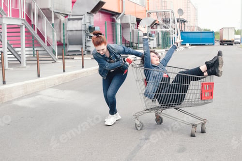 Preview: ✓ Funny happy teenage girls in denim clothes have fun and ride on the shopping cart