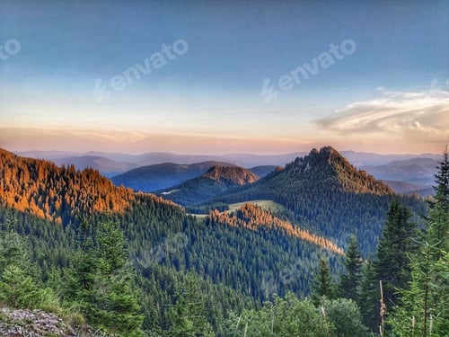 Preview: Aerial view of a mountain range covered with green forests under a cloudy blue sky at sunset