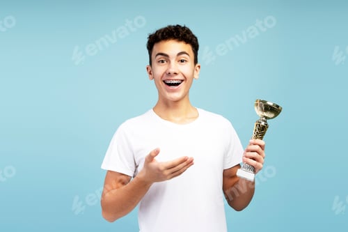 Preview: Excited boy with braces wearing white t shirt, holding trophy cup, pointing at it, looking at camera