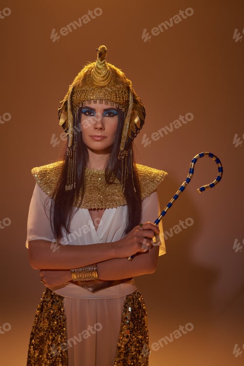 Preview: Brunette woman in egyptian attire and headdress holding crook and standing on brown background