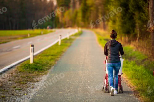 Vorschau: Junge Mutter und Kind im roten Kinderwagen gehen vor Sonnenuntergang im Freien in der ländlichen Gegend spazieren
