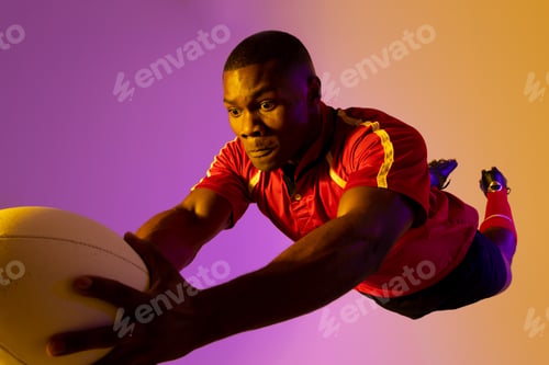 Preview: African american male rugby player jumping with rugby ball over pink lighting