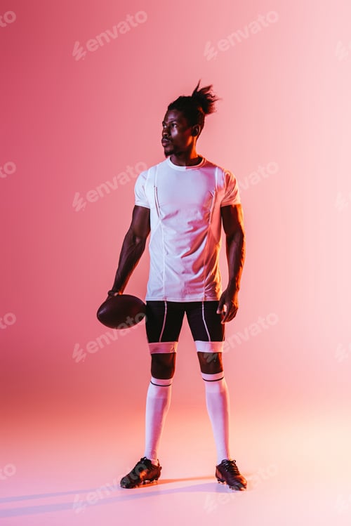 Preview: handsome african american sportsman looking away while holding rugby ball on pink background with