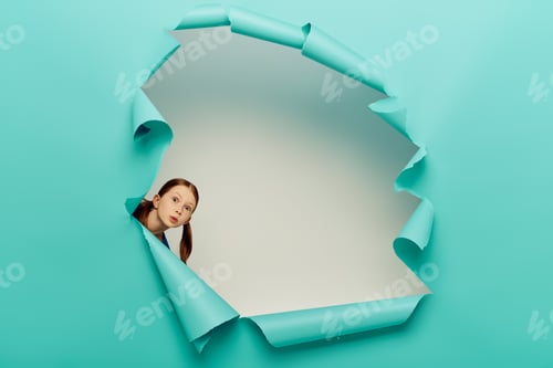 Preview: surprised redhead preteen girl looking at camera through blue torn paper hole on white background
