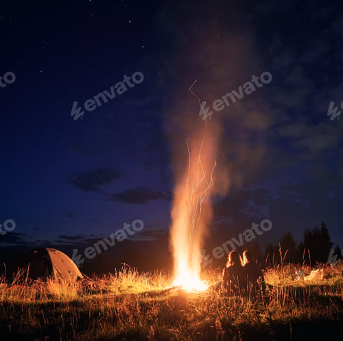 Preview: Young couple sitting near campfire under cloudy night sky.