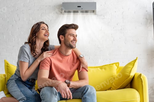 Preview: cheerful couple sitting on yellow sofa under air conditioner at home