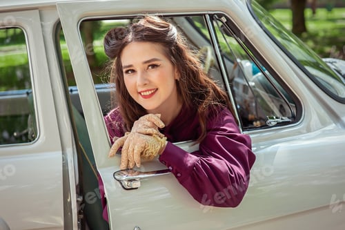Preview: Portrait of a beautiful smiling girl in a purple blouse looks out of the window of a retro car