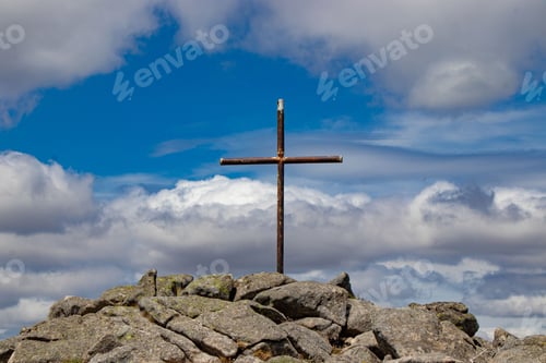 Preview: Wooden cross atop the peak of a rocky mountain against the cloudy blue sky