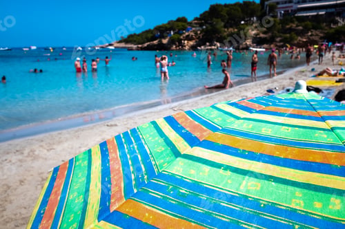 Preview: Vacationers enjoy a sunny day at the beach, background out of focus with parasol in the foreground.