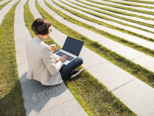 Preview: Business woman sits in park with laptop and take away cardboard cup of coffee.