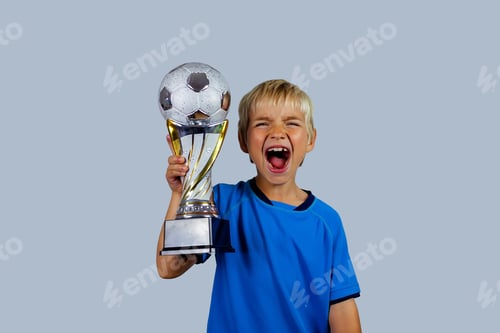Preview: Young soccer player in blue jersey holds winners cup after the goal, isolated at light background