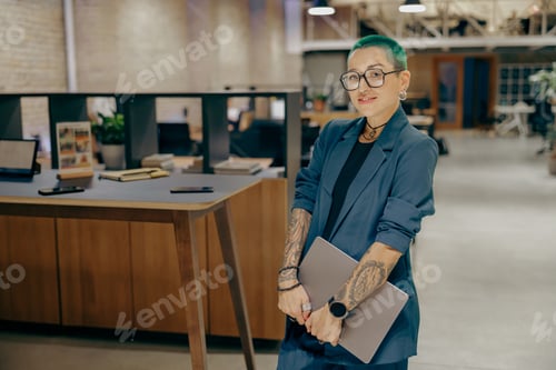 Preview: Smiling tattooed business woman with green short hair holding laptop standing in modern office