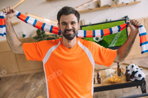 Preview: portrait of young man in orange fan t-shirt holding striped scarf and celebrating victory in soccer