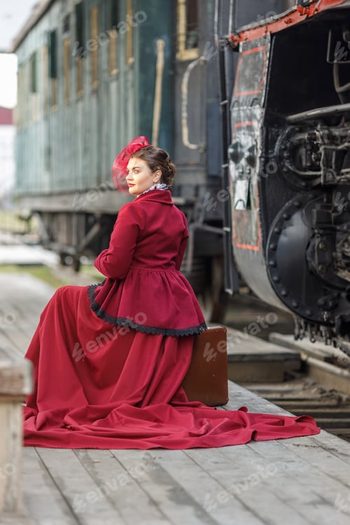 Preview: A beautiful girl in a burgundy suit is sitting on a suitcase near an old steam locomotive