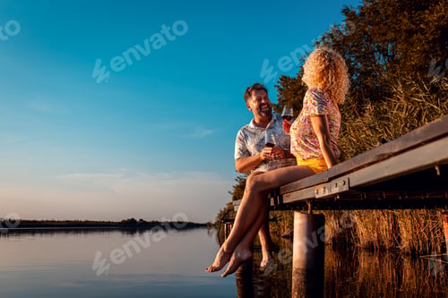 Preview: Smiling adult couple sitting by the lake on a pier drinking wine during sunset.