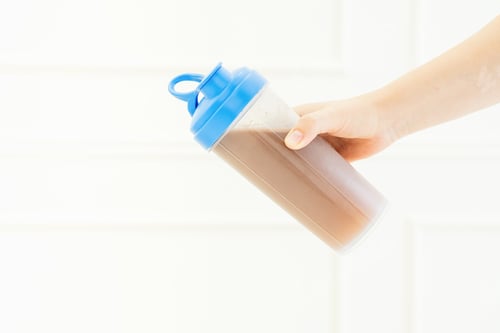 Preview: Female hand holding bottle with chocolate protein shake on white background close-up.