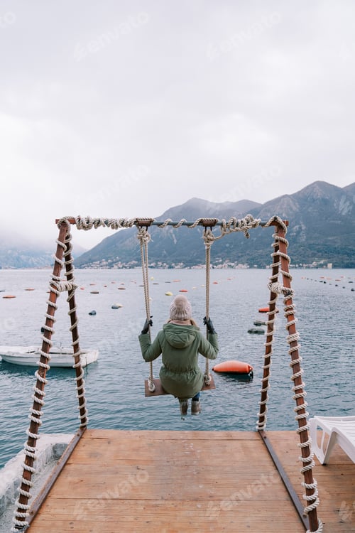 Preview: Girl in a jacket and hat swings on a swing on the pier and looks at the sea and mountains. Back view