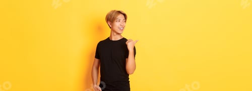Preview: Portrait of pleased thoughtful asian guy, looking and pointing finger left at logo, standing yellow