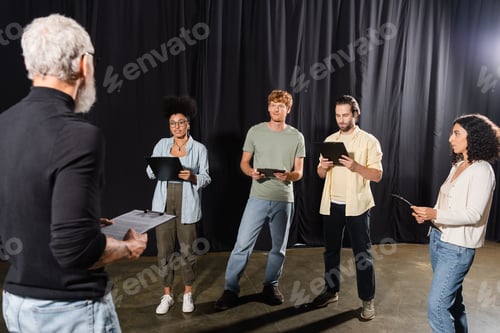 Preview: young interracial actors holding clipboards near grey haired screenwriter on blurred foreground