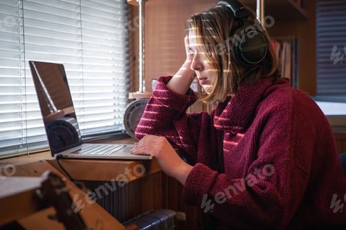 Preview: A girl with headphones behind a laptop in the sunlight, through the blinds.