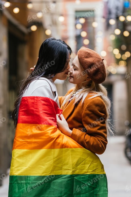 Preview: Lesbian couple kissing while wrapped in lgbt rainbow flag outdoors on the street.